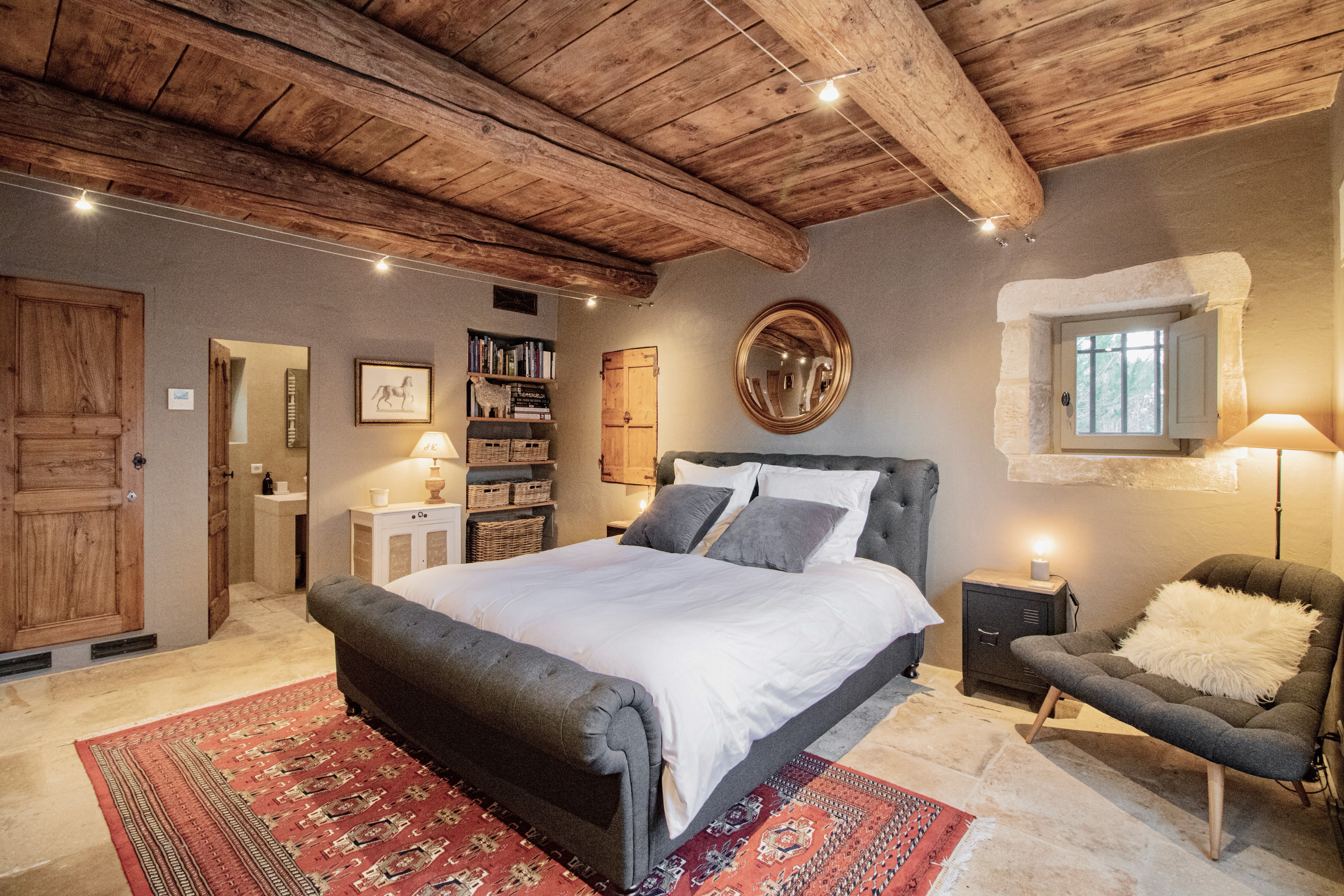 A rustic bedroom showcasing exposed wooden beams, a large gray bed, stone walls, a red patterned rug, wooden furniture, and a round mirror. The room has a cozy, warm ambiance.